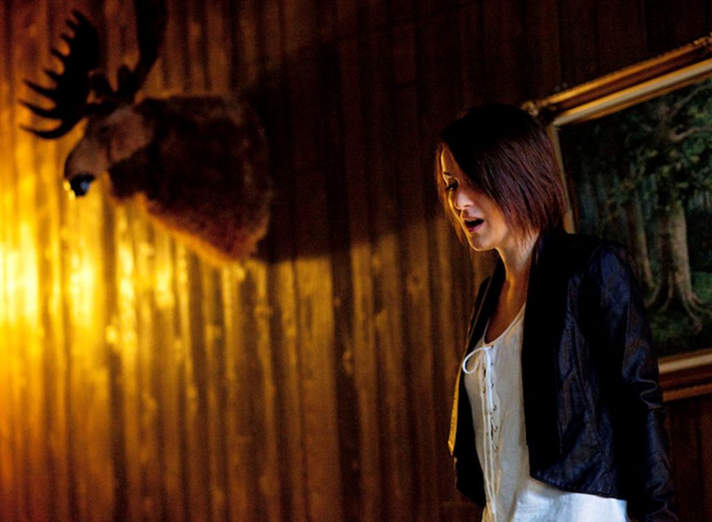 A woman in a black jacket and white shirt, is standing with her mouth open singing, there is a moose head on the wall behind her.