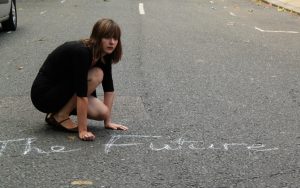 Deborah Pearson in a black dress is crouched on the road, next to the words 'The Future' written in chalk