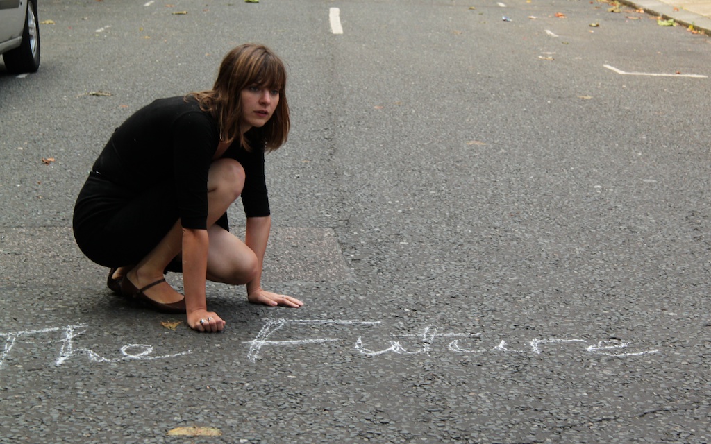 Deborah Pearson in a black dress is crouched on the road, next to the words 'The Future' written in chalk