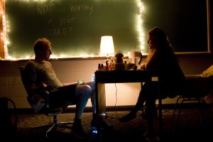 Two people are sat next to a table and a lamp talking to each other. In the background is a chalkboard with fairylights on it and the words 'What is Worthy of you CARE? in chalk.