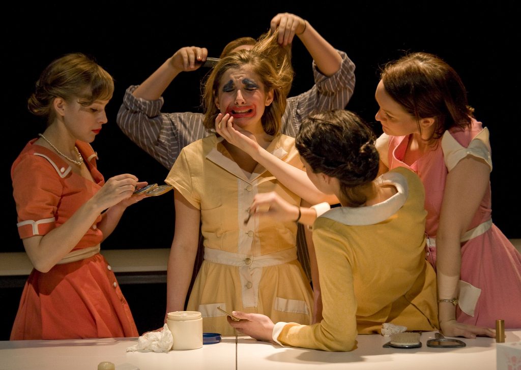 Four people are putting makeup on a crying central figure - the make up is like a clown. They are all dressed in fifties outfits