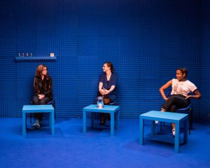 Three people sit in a blue soundproof room, each with a blue table in front of them. The middle table has a full jug of water on it.