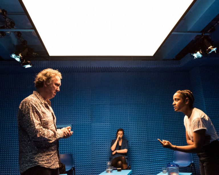 Two people standing opposite each other having a discussion in a blue soundproof room, between them another person sits with a blue table in front of them.
