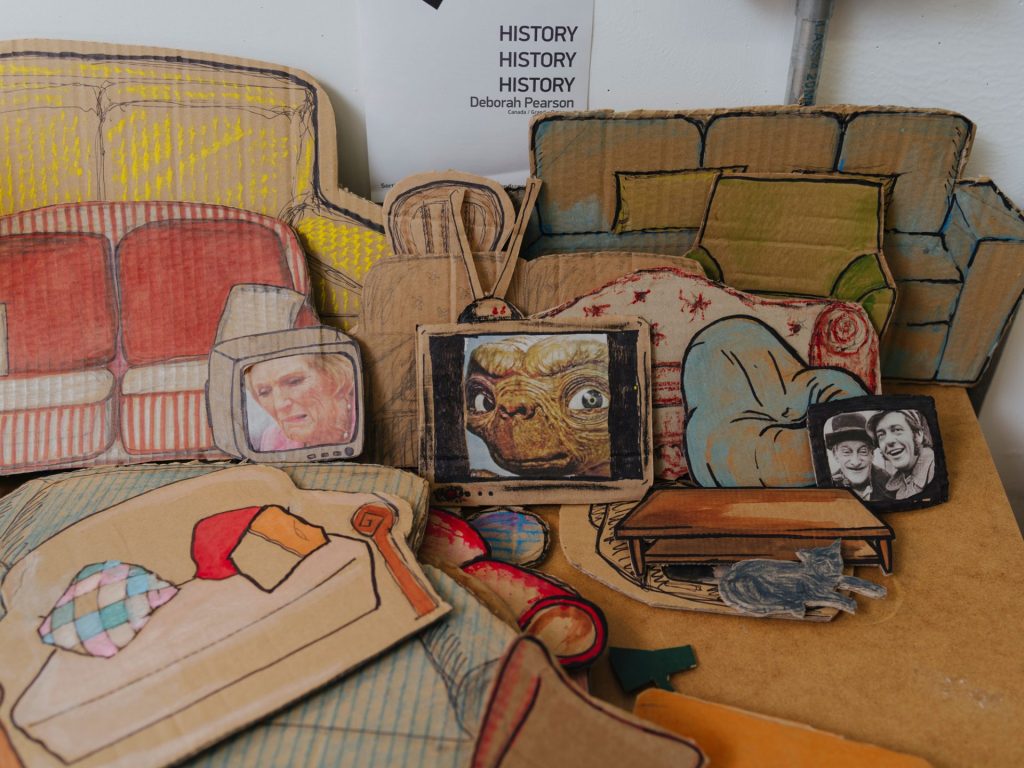 There is a pile of cardboard cut outs of furniture, such as tables, and sofas and TVs. On the wall behind is a sign that says 'History, History, History, Deborah Pearson, Canada)