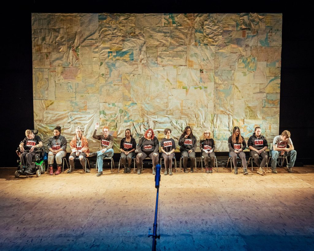 Twelve people each with a runners number pinned to their tops are sat on a stage. In the background there is a mosaic of maps. One person has their hands up. Everyone has their eyes shut.