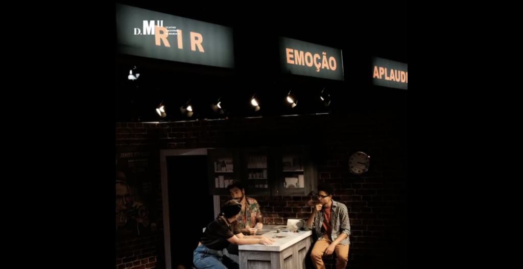 Three people are sat on a stage at a kitchen counter. Above them are signs - one which says 'Rir', one which says 'Emoção' and a thrid that says 'Aplaudio'