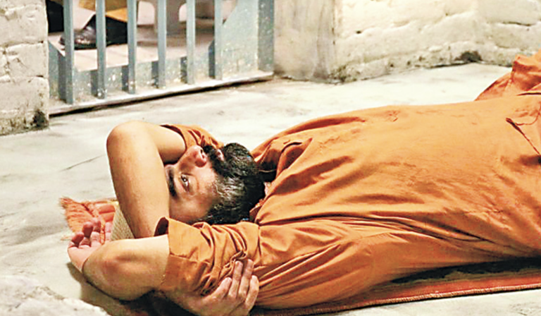 A man in an orange jumpsuit is lying on a cell on a rug.