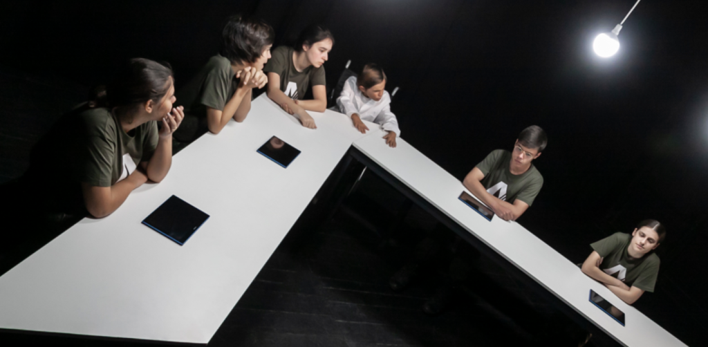 Around a long white table - six young people are sitting. They have tablets in front of them and a white lightbulb is brightly shining.