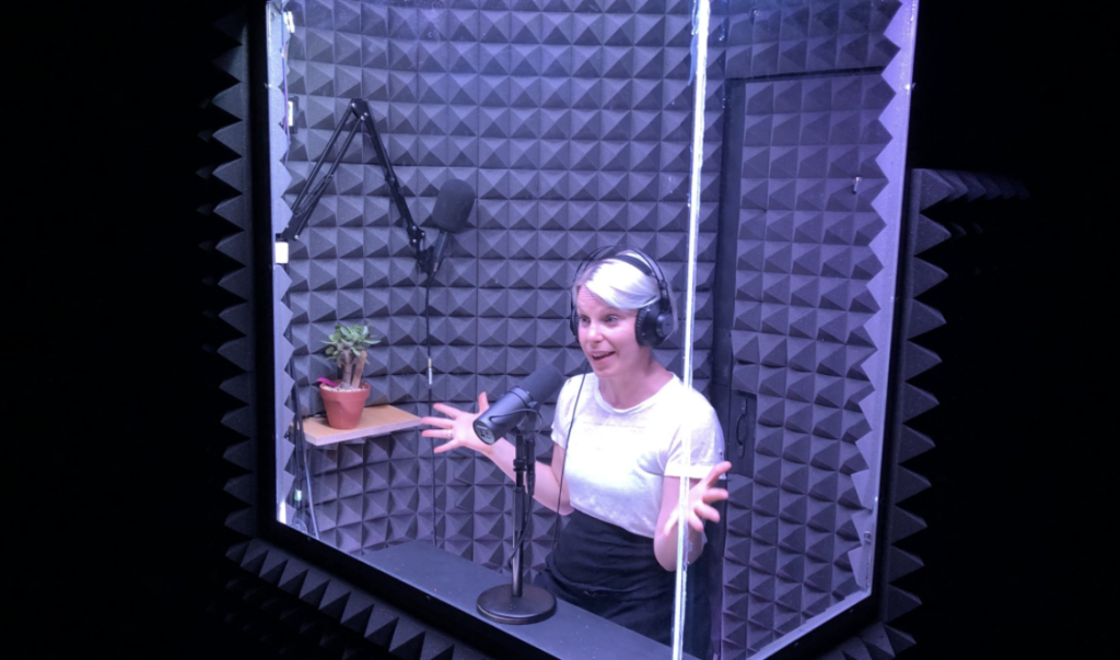 A woman is in a soundproof booth on a dark stage speaking into a microphone. She is wearing headphones smiling and has her hands wide stretched in a gesture.