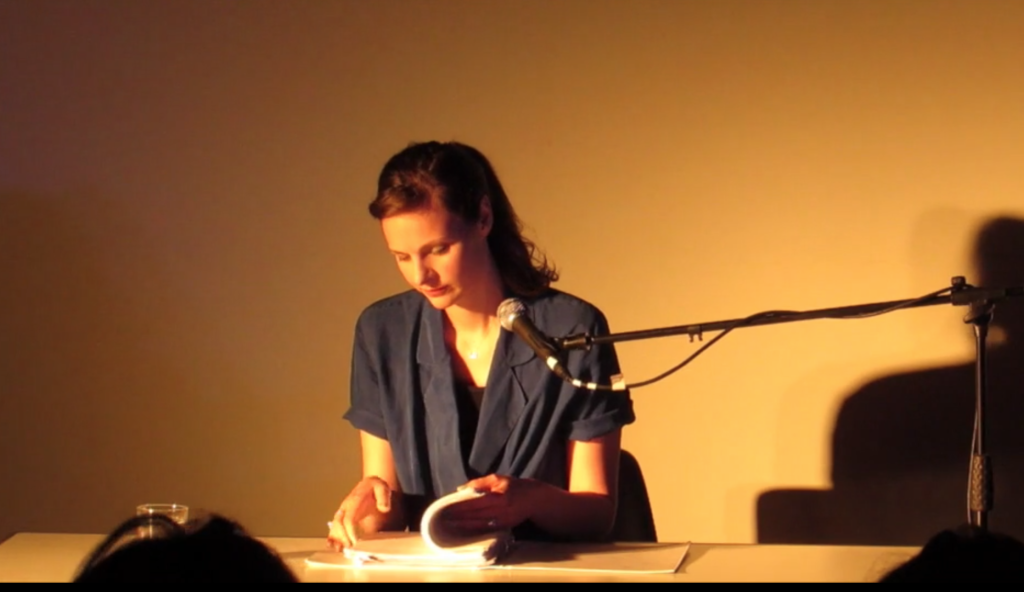 Deborah Pearson is sitting on stage at a desk, with a desk microphone and about to read from a text