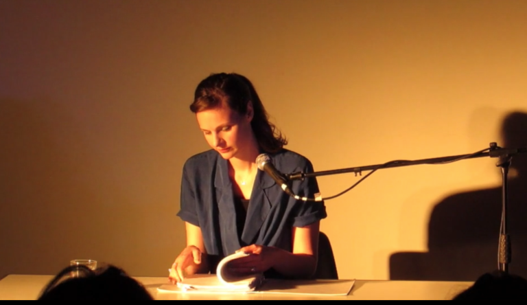 Deborah Pearson is sitting on stage at a desk, with a desk microphone and about to read from a text